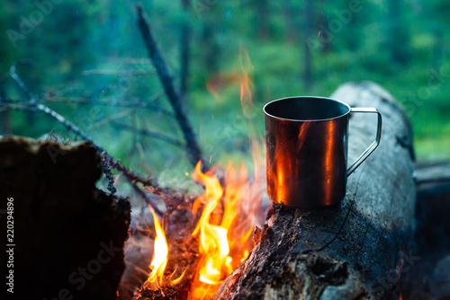 Tea in metal mug heats up on stone on bonfire. Hot drink on nature. Tea drinking in open air. Camping in dusk. Romantic warm atmosphere outdoor in twilight . Active rest.