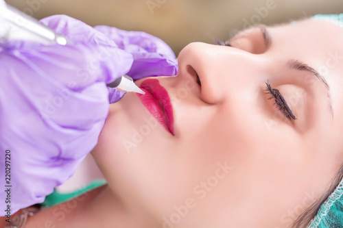 Young woman having permanent makeup on lips in beautician salon. Cosmetologist applying tattoo with professional tool. Close up, selective focus