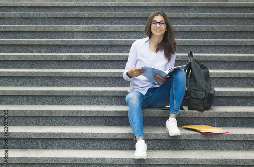 Beautiful smiling student girl with textbooks, folders, near college. A young Indian woman in glasses sits on the steps and reads books. 