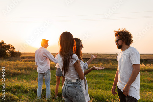 Two young dark-haired women chatting with young curly man and laughing outdoor on a warm day.