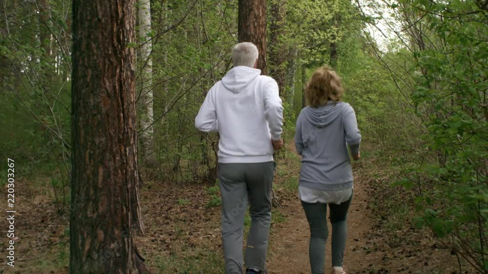 Dolly shot with rear view of elderly man and woman running along path in forest Dolly shot with rear view of elderly man and woman running along path in forest