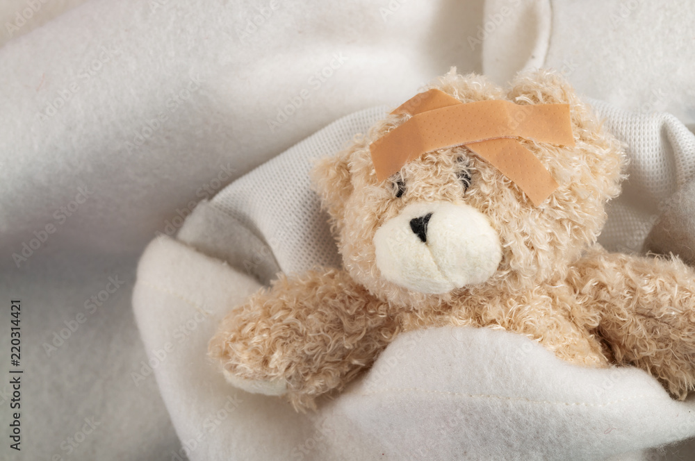 Childhood illness and sick kids concept with a ill teddy bear covered in a blanket with a adhesive bandages on its head to cover an injury with copy space