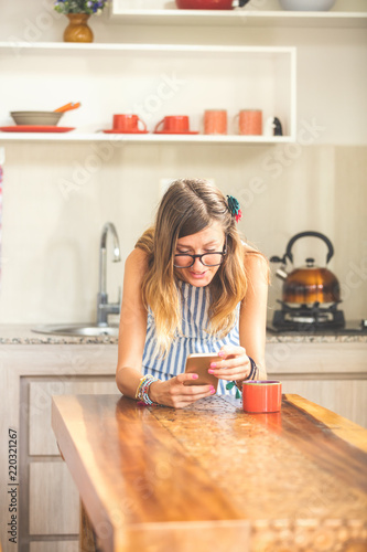Young woman using cellphone and drinking coffee / tea in a modern kitchen.