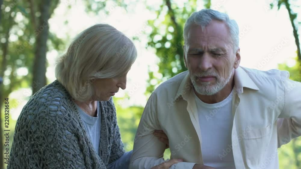 Mature man feeling sharp back pain while walking in park caring wife helping him Mature man feeling sharp back pain while walking in park caring wife helping him