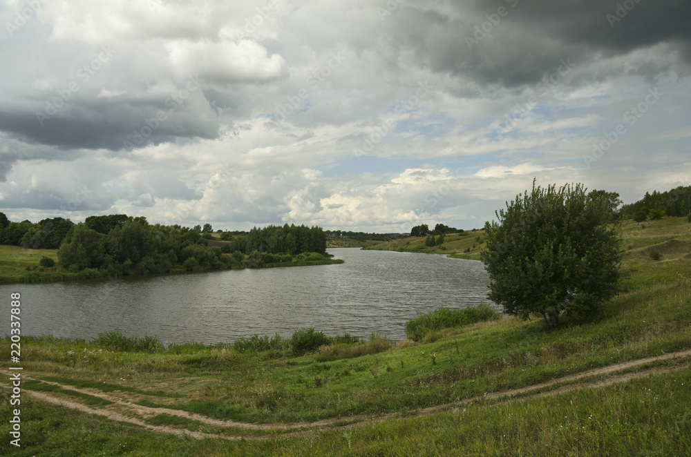 Fototapeta premium Cloudy summer landscape with small river.Dark stormy clouds in dramatic overcast sky.Fields,green meadows and woods.Rainy season