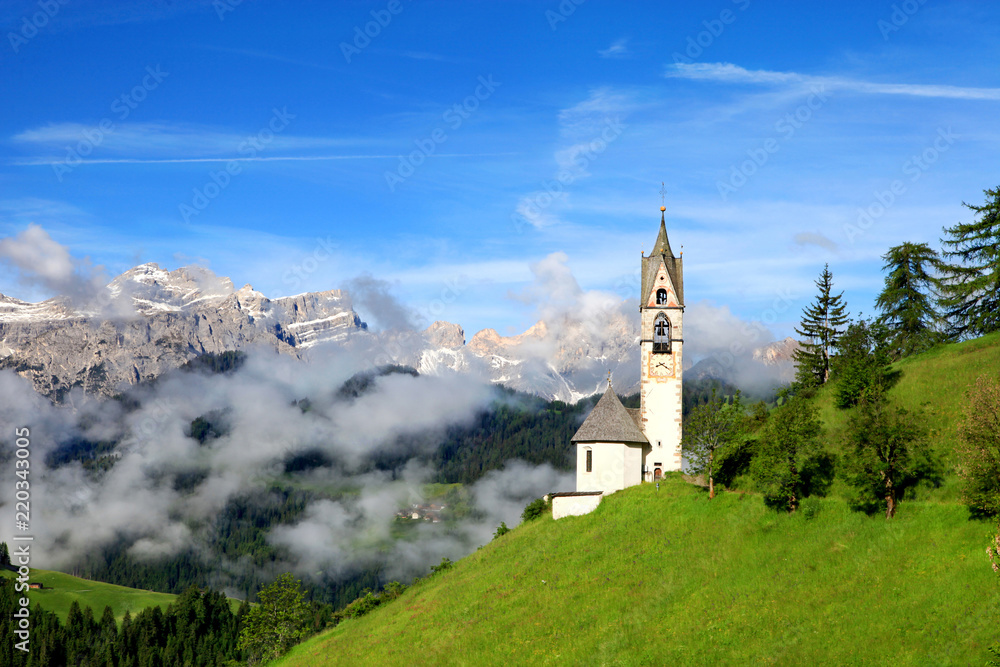 Santa Barbara Church in the early morning in the foggy valley with clouds covered the mountain. The chapel is on a hill, offering to its visitors a panoramic view in La Valle, Dolomites, Italy.