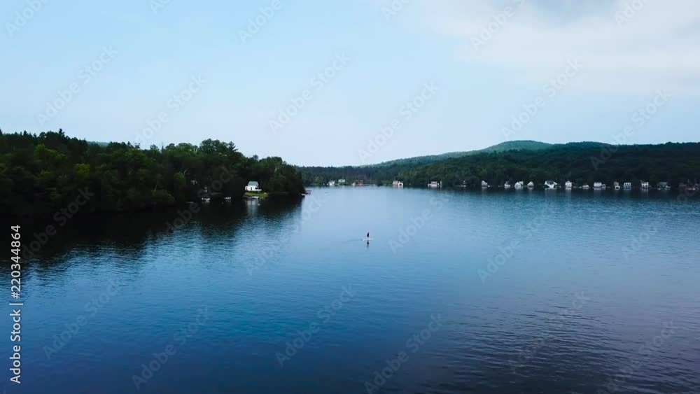 A women paddles out on the lake. The drone starts off as a wide shot and quickly zooms in getting a close shot of the paddle boarder. It moves past the boarder and towards the center of the lake. A women paddles out on the lake. The drone starts off as a wide shot and quickly zooms in getting a close shot of the paddle boarder. It moves past the boarder and towards the center of the lake.