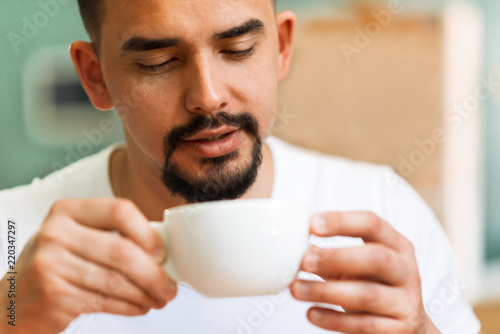 Tanned man with beard and moustache keeping with both hands a white mug of a hot coffee and eagerly looking at it with love