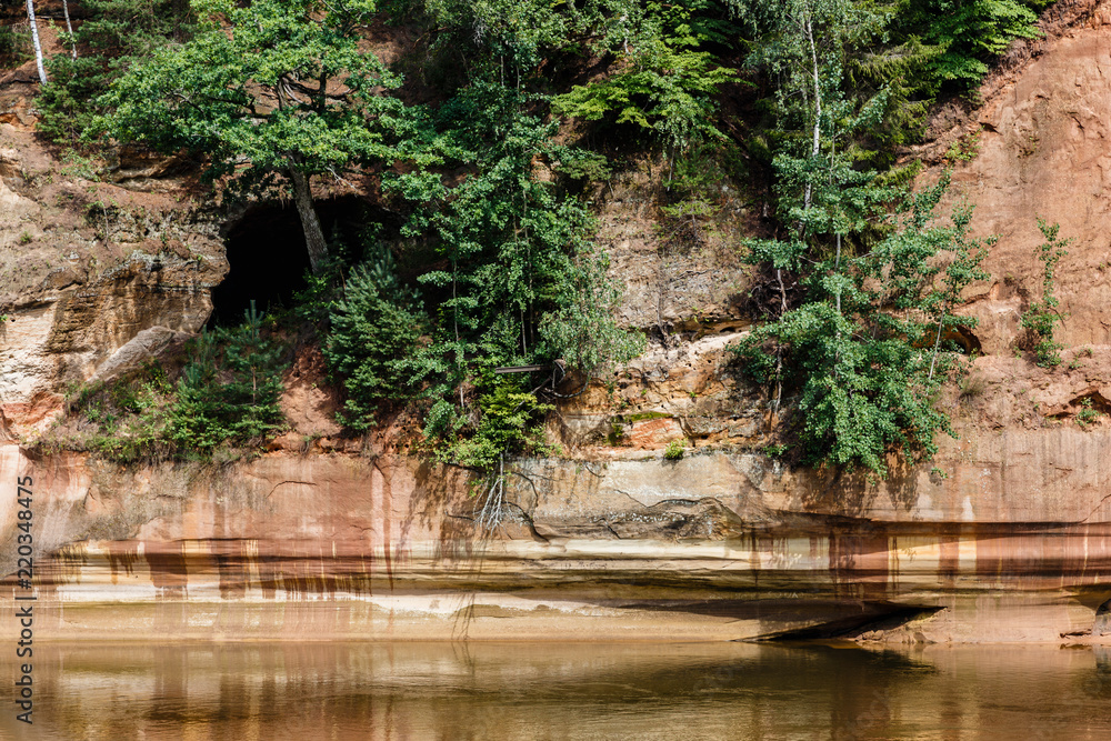 Fototapeta premium sunny summer day at the river and the red rocks of the cliff grow some trees, rock reflection in the river