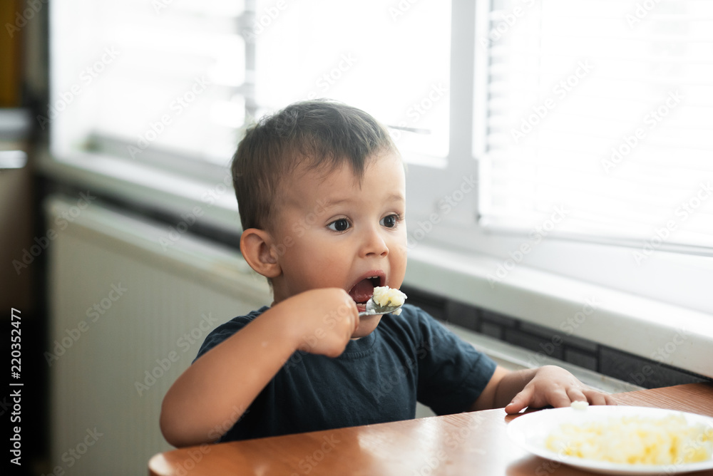The little boy in the kitchen eagerly eating rice with a spoon independently The little boy in the kitchen eagerly eating rice with a spoon independently