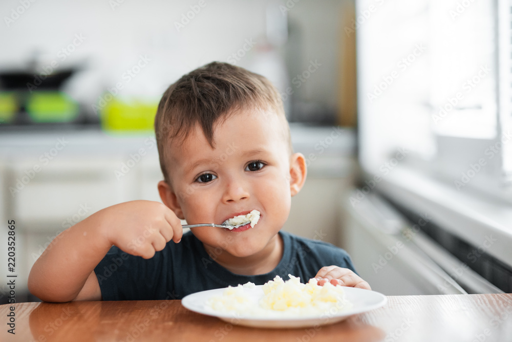 The little boy in the kitchen eagerly eating rice with a spoon independently The little boy in the kitchen eagerly eating rice with a spoon independently