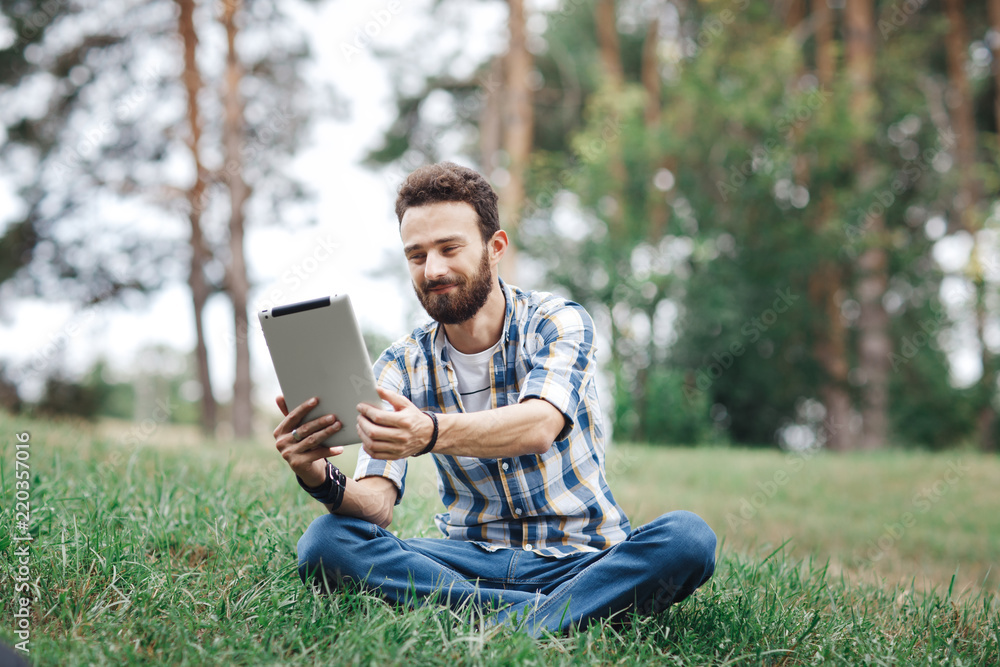 Smiling young bearded hipster sitting on the grass. Bearded man with tablet and headphones listening music in the park. Smiling young bearded hipster sitting on the grass. Bearded man with tablet and headphones listening music in the park.