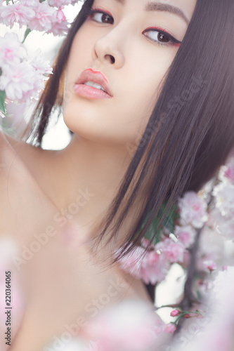 Outdoor fashion photo of beautiful young asian woman surrounded by flowers on spring. Perfect model with creative vivid makeup and pink lipstick on lips and traditional japanese hairstyle posing