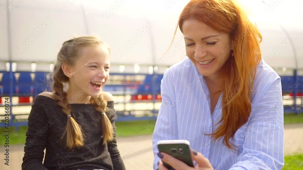 young mom and her daughter are watching a hilarious smartphone video and smiling while sitting in the Park for a walk