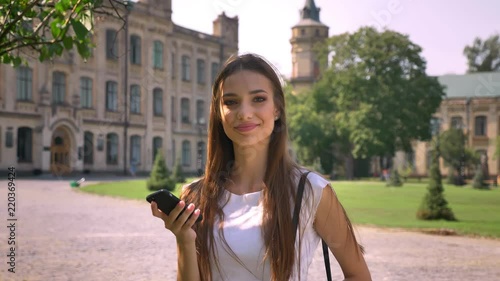 Sexy lady is standing in park in daytime, watchimg in smartphone then at camera photo