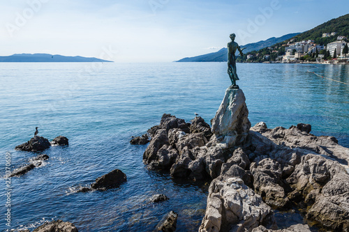 seaside village near the Mediterranean Sea, on the rocky side of the sea there is a monument - a girl with a bird on her hands; beautiful blue waters and mountains in the distance