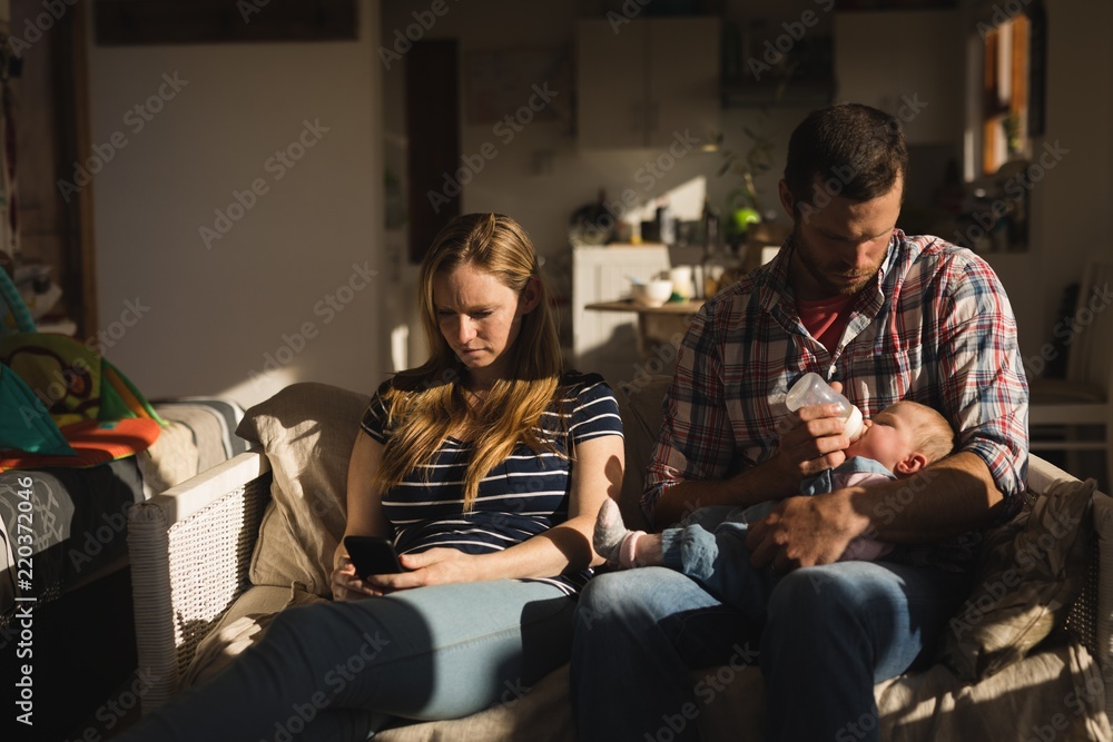 Mother using mobile phone while father feeding his baby on sofa Mother using mobile phone while father feeding his baby on sofa