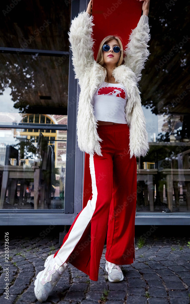 Fashion young girl posing in blue sunglasses and white fur. Stylish woman in in white fur and sport red pants and sneakers .Motion shot.