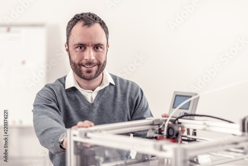 Interesting job. Pleasant bearded worker smiling standing near 3d printer working with it