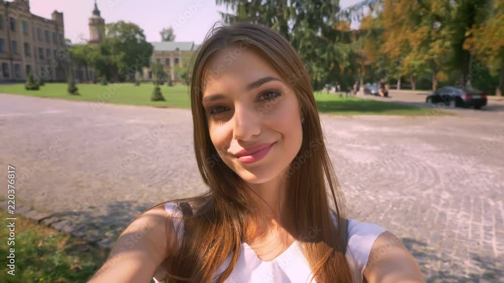 Nice young woman is standing in park in daytime, holds camera, makes selfie, smiling, communication concept Nice young woman is standing in park in daytime, holds camera, makes selfie, smiling, communication concept