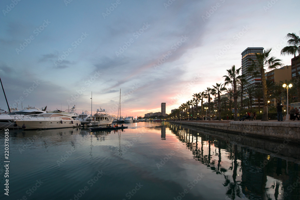 Beautiful port of Alicante, Spain at Mediterranean sea. Luxury yachts, ships, ferries and fishing boats sailing and standing in rows in harbor. Rich people traveling around the world. Sunset evening