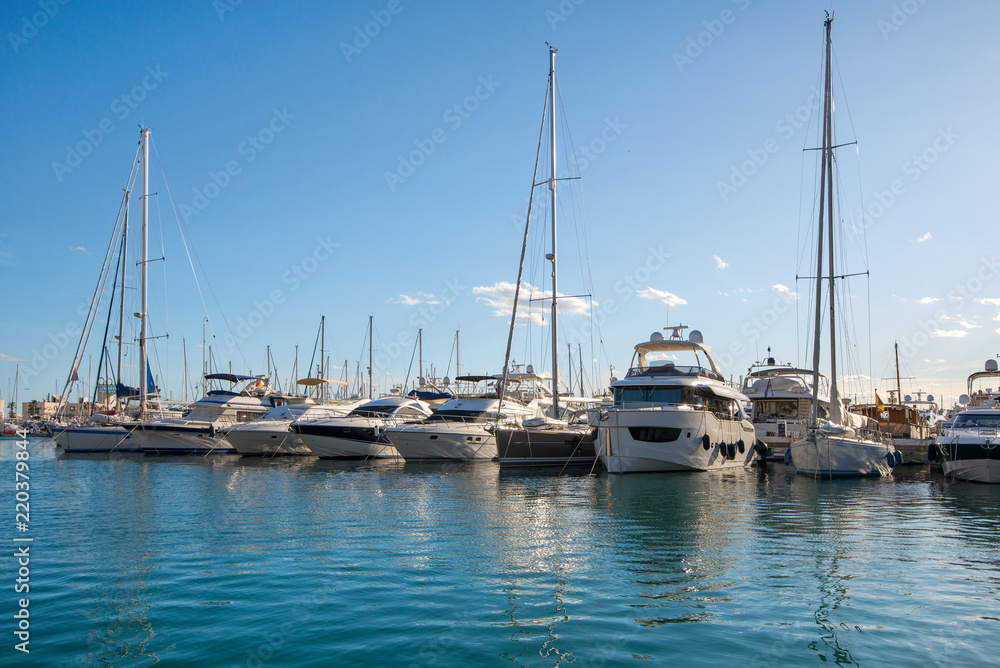 Beautiful port of Alicante, Spain at Mediterranean sea. Luxury yachts, ships, ferries and fishing boats sailing and standing in rows in harbor. Rich people traveling around the world. Sunny evening