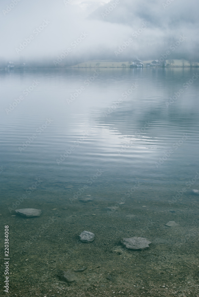 Mysterious mountain lake in early spring morning. Spooky dense fog and bright morning light reflected in water. Sand bottom covered with stones Mysterious mountain lake in early spring morning. Spooky dense fog and bright morning light reflected in water. Sand bottom covered with stones