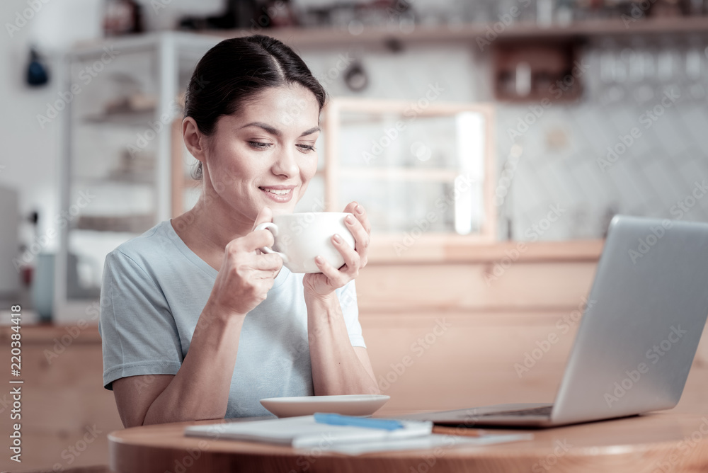 Delicious smell. Calm young woman relaxing in a cafe and holding a cup of delicious coffee while sitting in front of a modern laptop Delicious smell. Calm young woman relaxing in a cafe and holding a cup of delicious coffee while sitting in front of a modern laptop