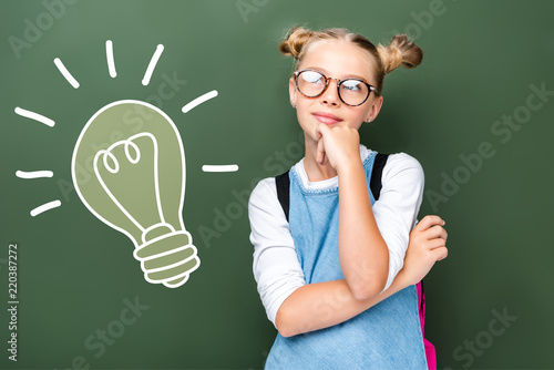 pensive schoolchild in glasses looking up near blackboard with light bulb sign
