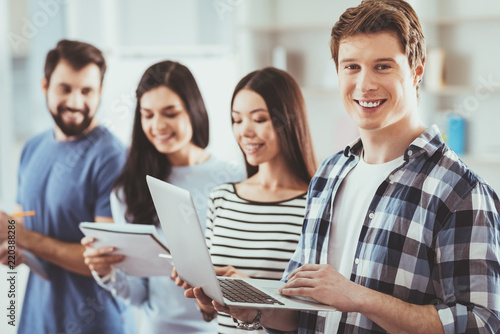 Team leadership. Delighted positive man smiling to you while holding his laptop