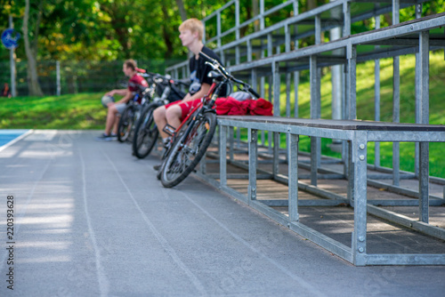 Cyclists resting and sitting on metal bench next to outdoor basketball court and watching the game.  Healthy lifestyle and outdoor activities in  summer, spring and autumn © lainen