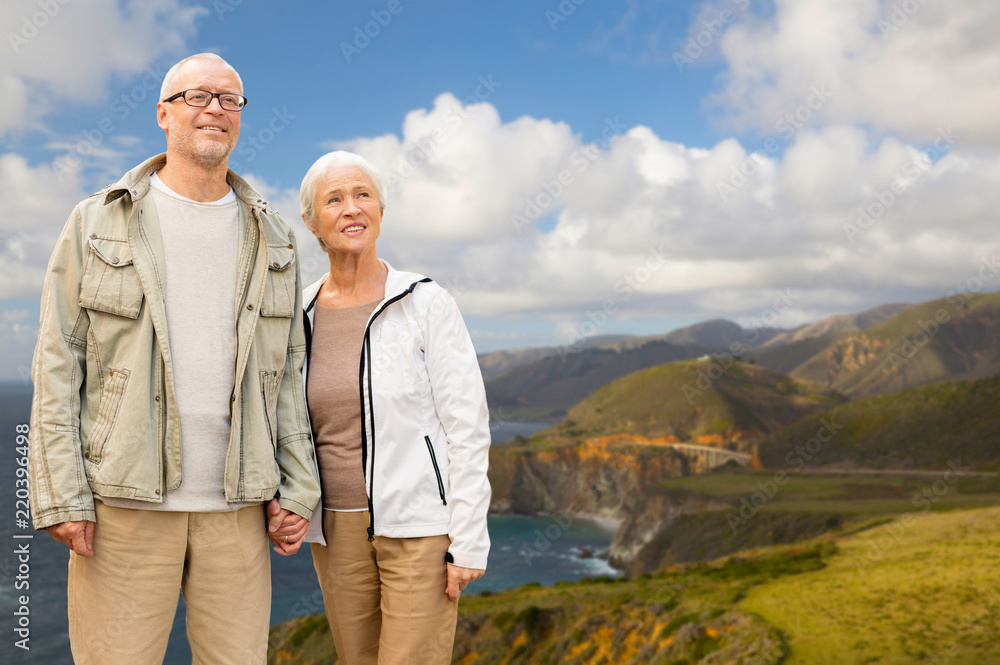 old age, tourism, travel and people concept - happy senior couple holding hands over bixby creek bridge on big sur coast of california background
