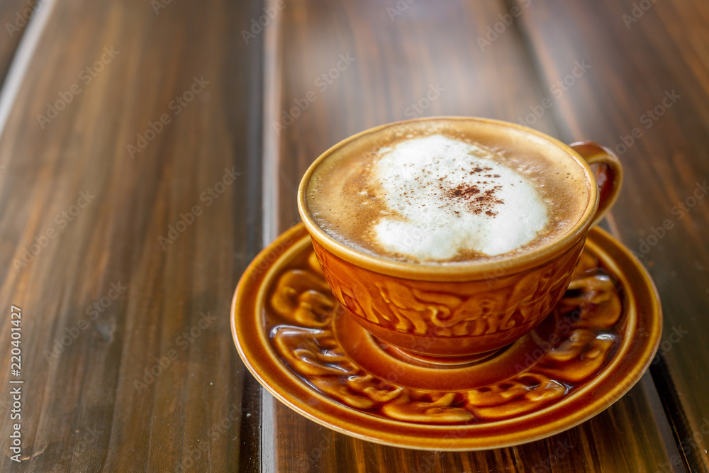 The white ceramic cup of hot capucchino coffee on the wooden table in the restaurant or coffee shop with blur people at the background in thw warm yellow light and flare. The white ceramic cup of hot capucchino coffee on the wooden table in the restaurant or coffee shop with blur people at the background in thw warm yellow light and flare.