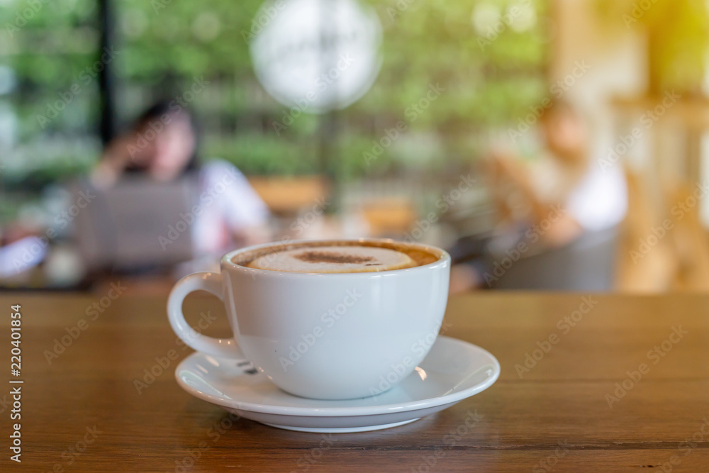 The  white ceramic cup of hot capucchino coffee on the wooden table in the restaurant or coffee shop with blur people at the background in thw warm yellow light and flare. 