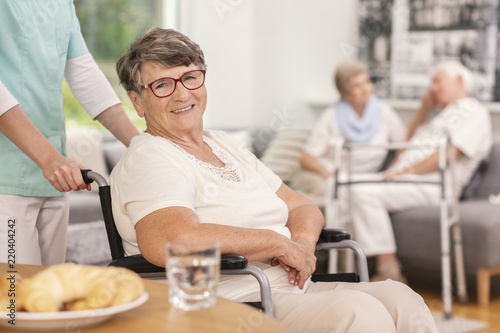 A geriatric female patient in a wheelchair with a health assistant in a lobby of a luxurious private rehabilitation clinic. Blurred background.