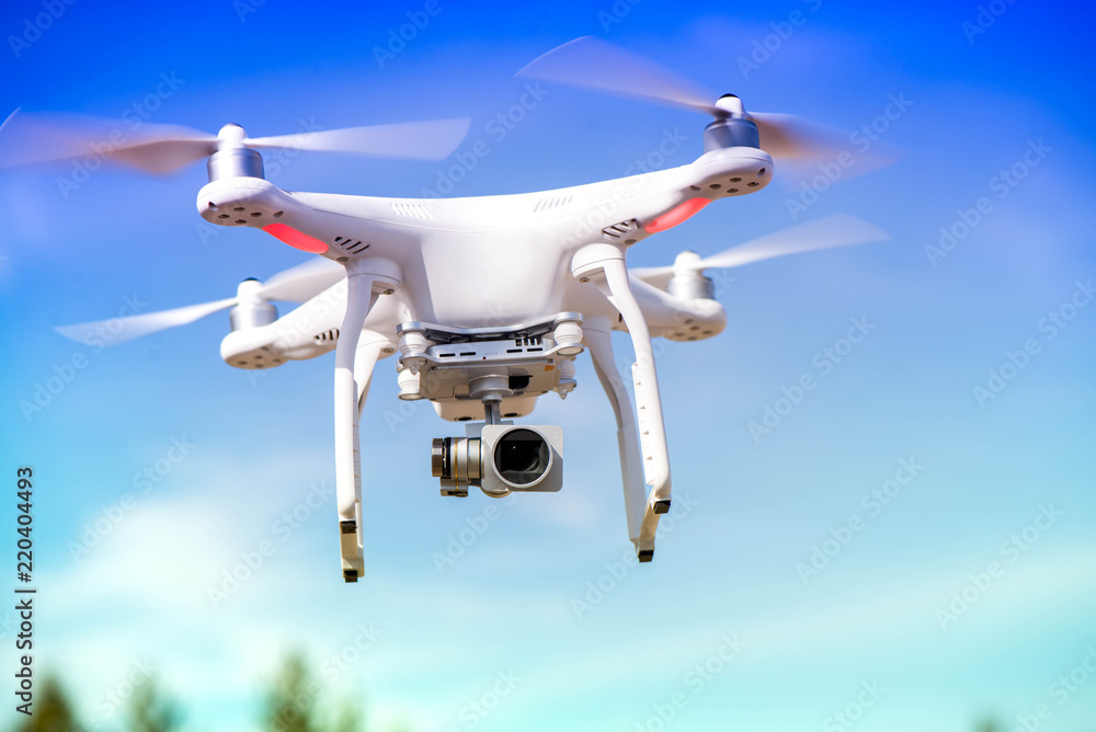 White quadrocopter is flying high in the air, taking photos and recording footage from above. Flying drone with four motors and propellers, camera and red warning lights on clear blue sky background - obrazy, fototapety, plakaty 