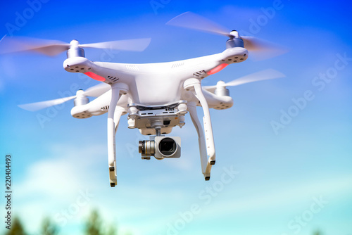 White quadrocopter is flying high in the air, taking photos and recording footage from above. Flying drone with four motors and propellers, camera and red warning lights on clear blue sky background
