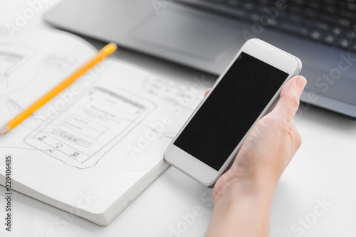 app design, technology and business concept - hand of web designer or developer with smartphone and sketch of user interface in notebook at office
