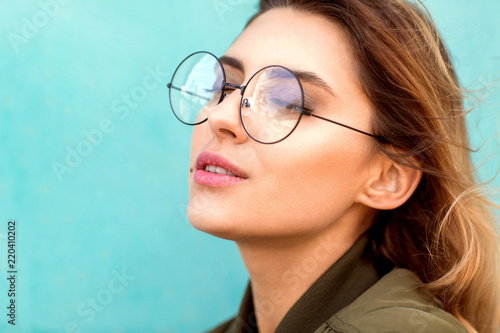 fashion girl in round glasses stands posing near a turquoise wall