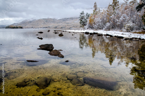 Mountain lake Froliha, pine tree and stones via transparent water with snow at mirror lake