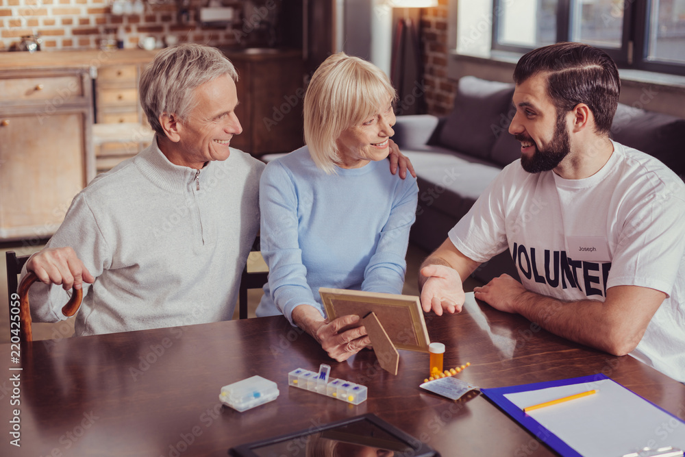 Tell me about it. Attentive cute senior woman sitting with her husband by the table holding a photo and looking at a volunteer. Tell me about it. Attentive cute senior woman sitting with her husband by the table holding a photo and looking at a volunteer.
