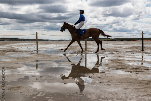 Reflection in a water pool of race horse and jockey galloping on the beach
