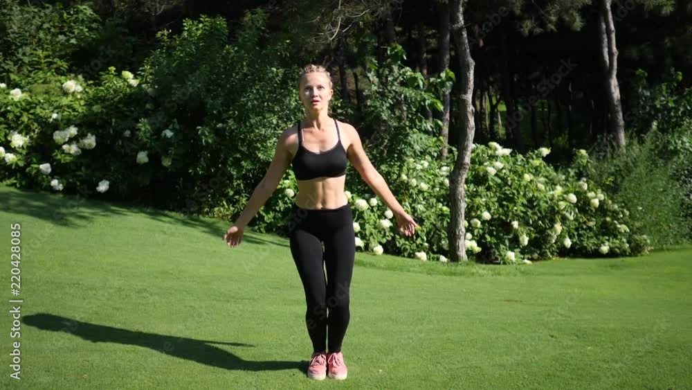 Young blond woman with braids doing jumping jacks fitness exercise workout on the meadow in the green summer park. Slow motion Young blond woman with braids doing jumping jacks fitness exercise workout on the meadow in the green summer park. Slow motion