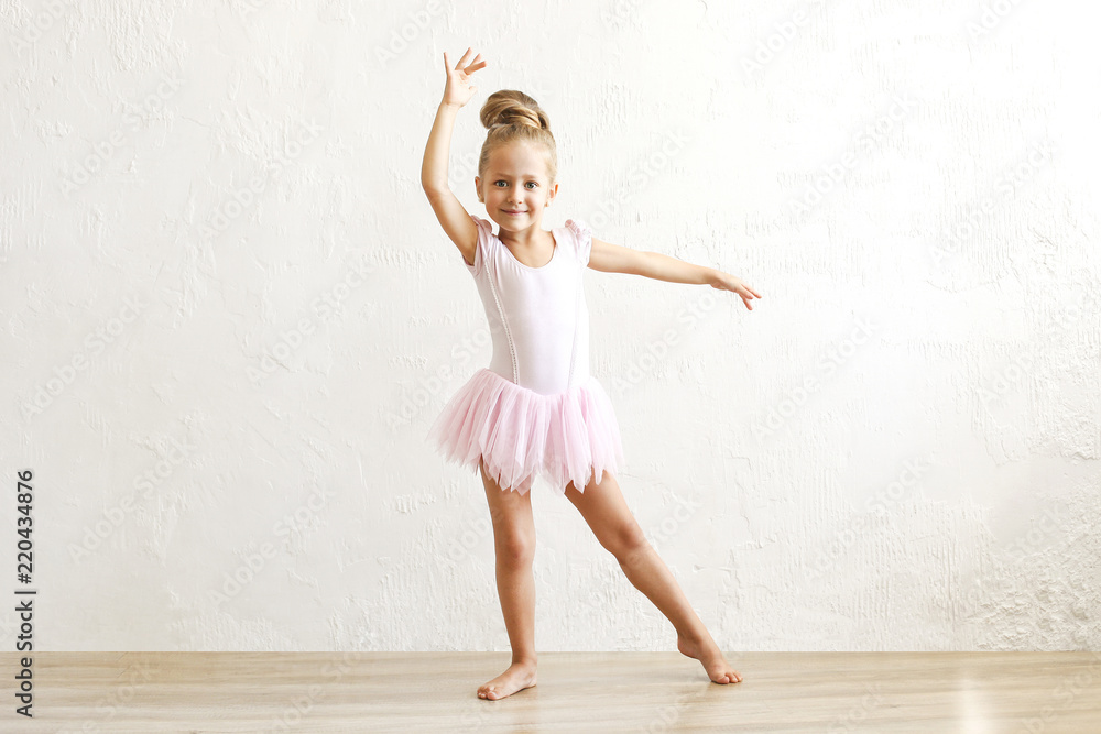 Little blonde balerina girl dancing and posing in dance club with wooden  floot an white textured
