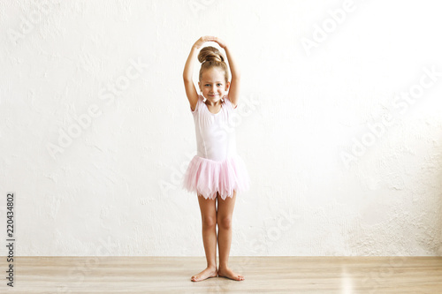 Little blonde balerina girl dancing and posing in dance club with wooden floot an white textured plaster wall. Young ballet dancer in pink tutu dress, having fun and smiling. Backgroud, copy space. photo