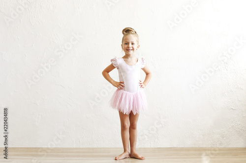Little blonde balerina girl dancing and posing in dance club with wooden floot an white textured plaster wall. Young ballet dancer in pink tutu dress, having fun and smiling. Backgroud, copy space.
