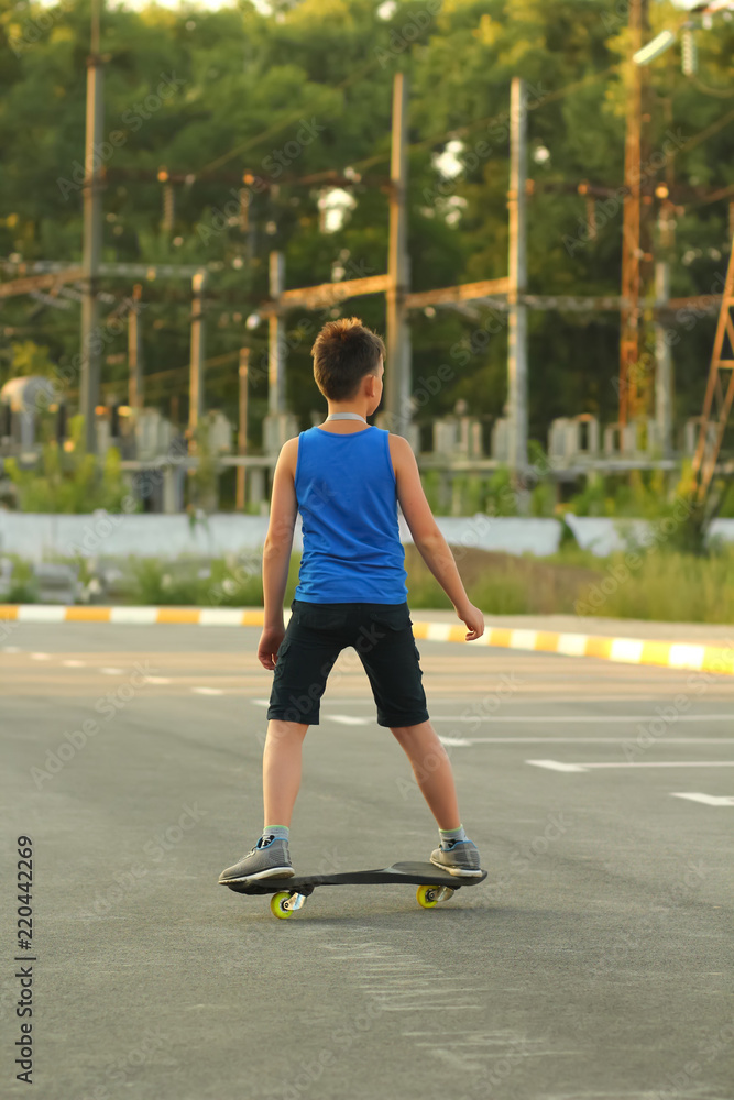 Boy riding skate board on summer parking lot near power station, back view