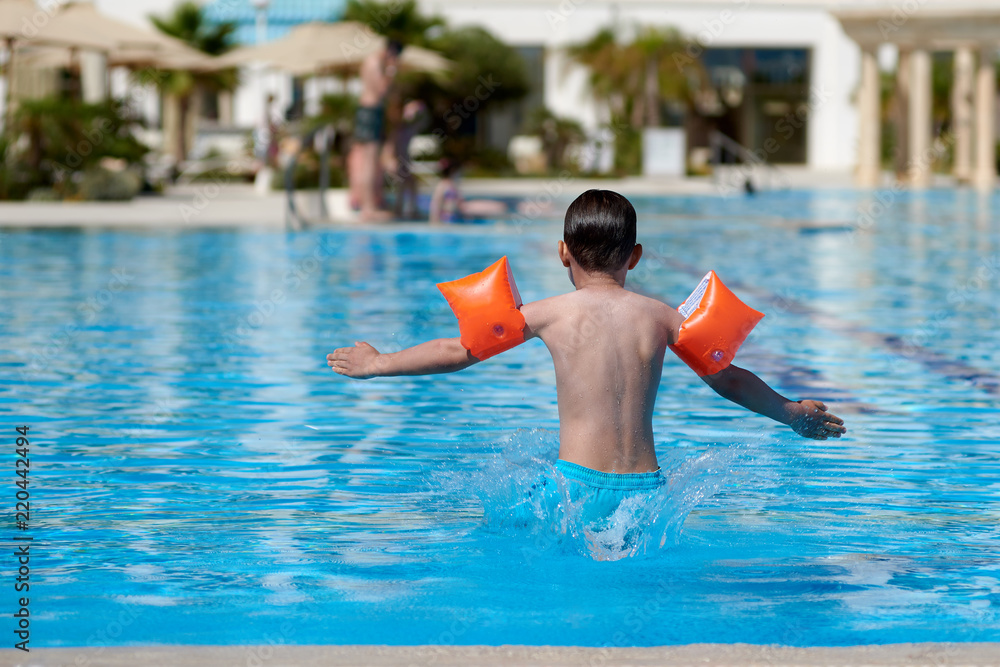 European boy in floating sleeves jumping into swimming pool at resort. Moment of entrance in water.