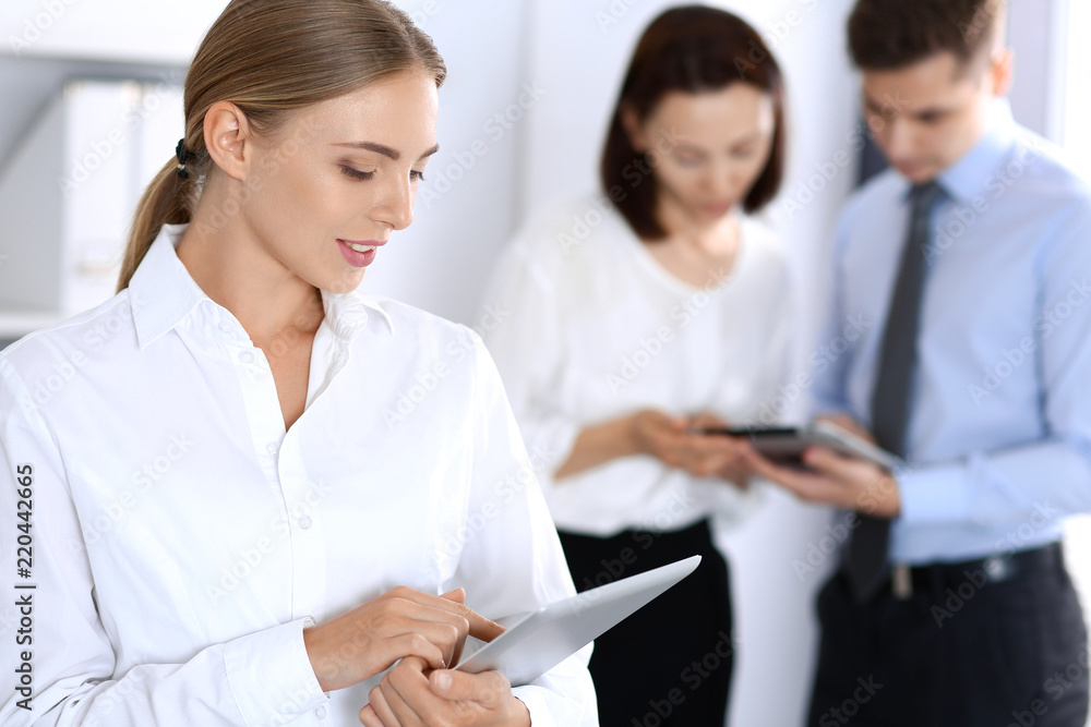 Beautiful blonde business woman using tablet or touch-pad on background of colleagues in office Beautiful blonde business woman using tablet or touch-pad on background of colleagues in office