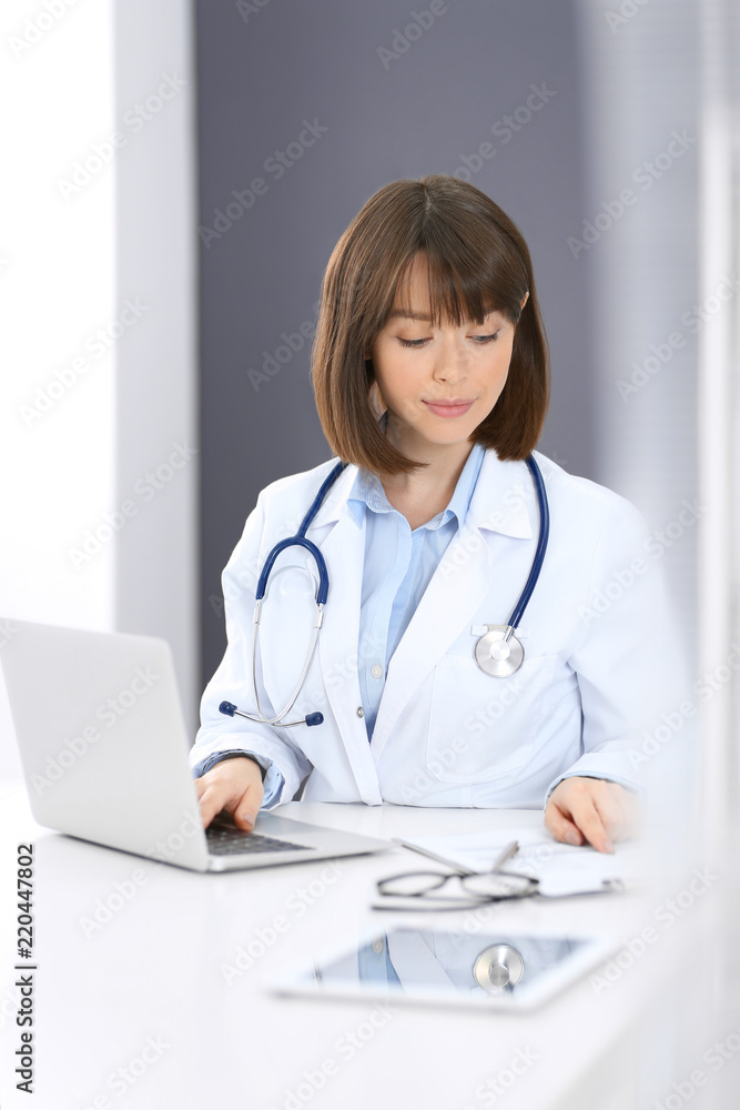 Doctor typing on laptop computer while sitting at the white table in hospital office. Physician at work. Medicine and healthcare concept Doctor typing on laptop computer while sitting at the white table in hospital office. Physician at work. Medicine and healthcare concept